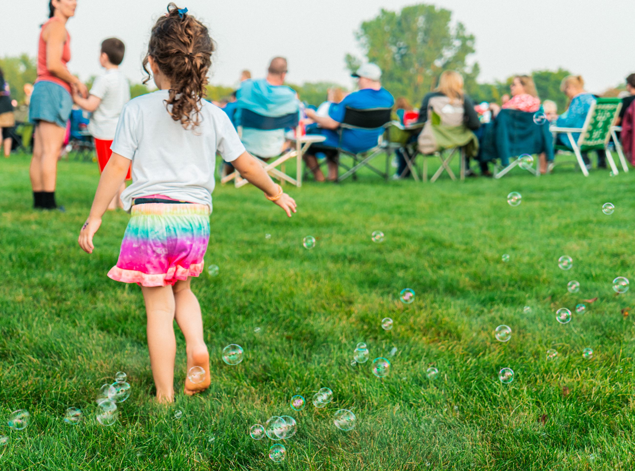 A little girl walks barefoot in the grass through bubbles towards people in lawn chairs